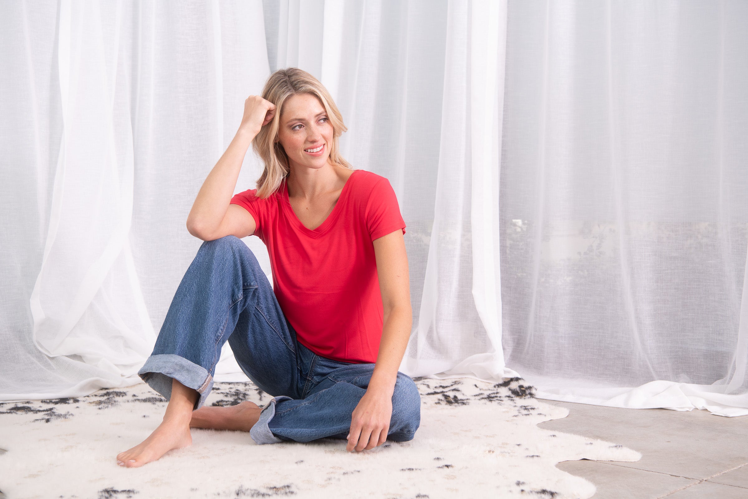 A woman with blonde hair sits cross-legged on a white rug in front of white sheer curtains, wearing a red short-sleeve t-shirt and blue jeans.