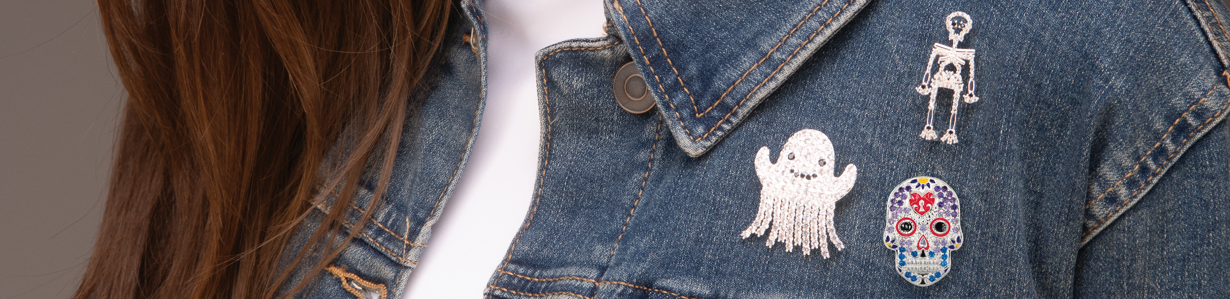 Close-up of a denim jacket worn by a person, featuring three embroidered Halloween-themed pins: a white ghost, a silver skeleton, and a sugar skull with red and blue details.