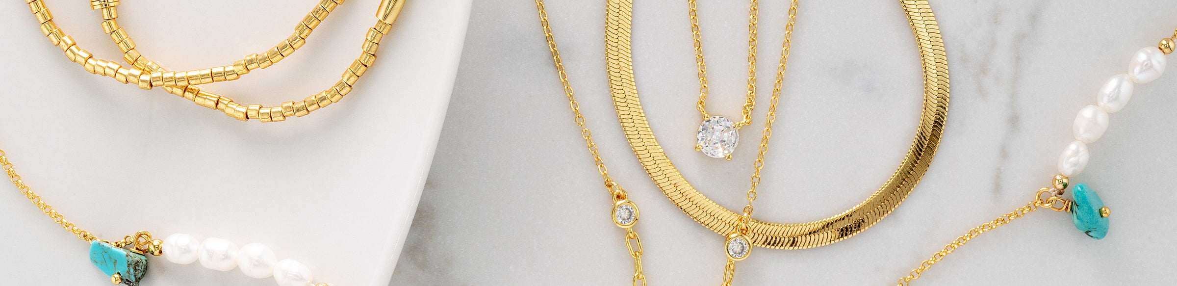 A close-up of layered gold necklaces displayed on a white marble surface, including a snake chain, a beaded pearl-and-gold necklace, and a chain with turquoise and clear stone accents.