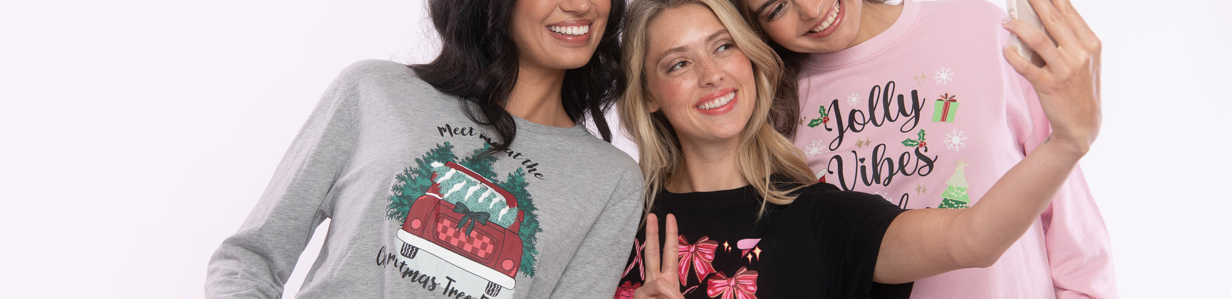 Three women posing together and taking a selfie while wearing matching long-sleeve holiday graphic sweatshirts in gray, black, and pink. The sweatshirts feature festive Christmas-themed graphics and text including 'Jolly Vibes' and 'Winter Trees'.