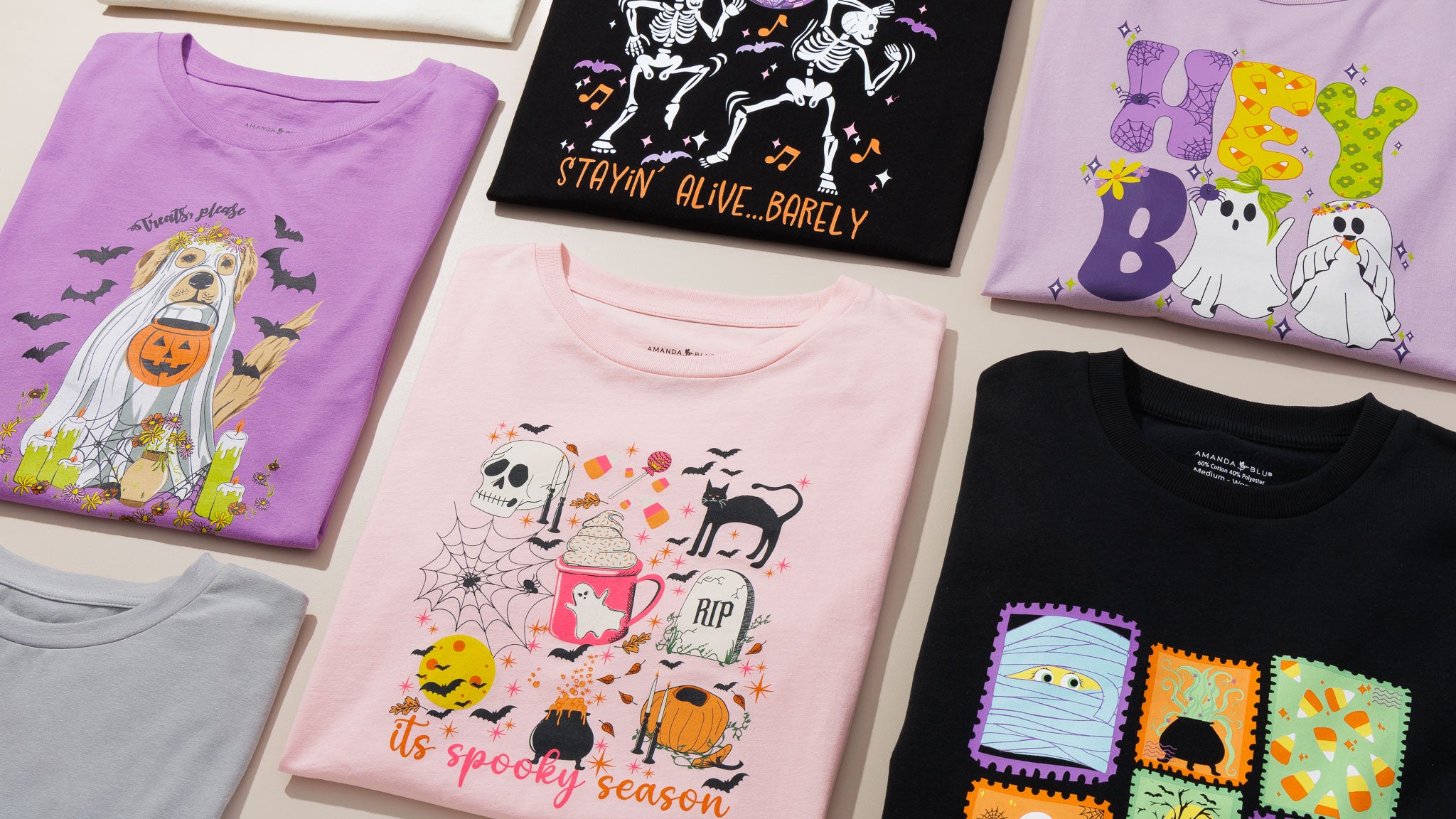 A flat-lay display of several folded graphic T-shirts with Halloween-themed designs, including a purple dog in a ghost costume, a black skeleton bandit shirt, a pink spooky mugshot shirt, and other partially visible shirts.