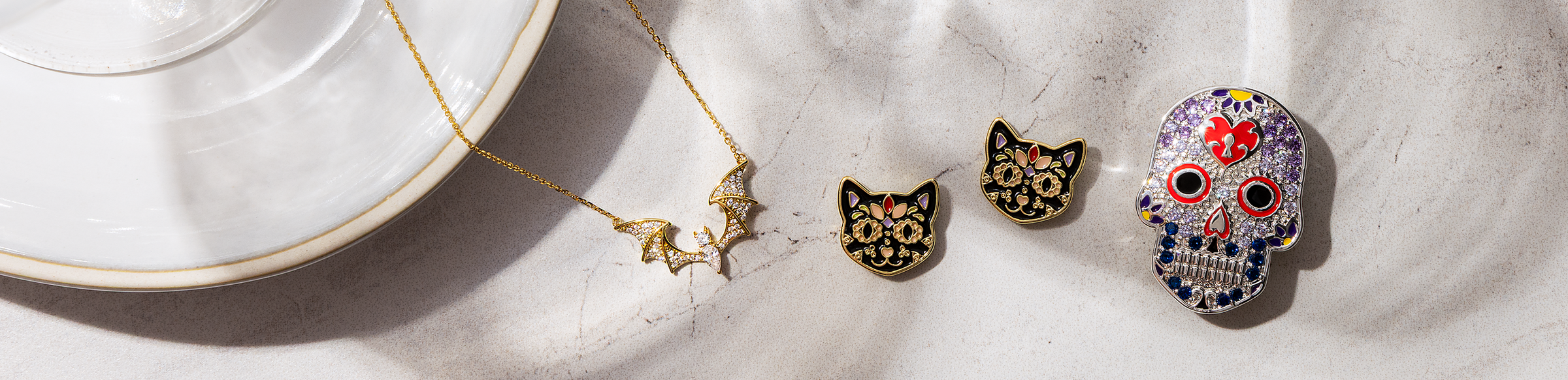 A styled flat-lay of jewelry on a light textured fabric next to a white plate, featuring a gold necklace with a butterfly, two black cat-shaped earrings with gold floral details, and a colorful sugar skull pendant.
