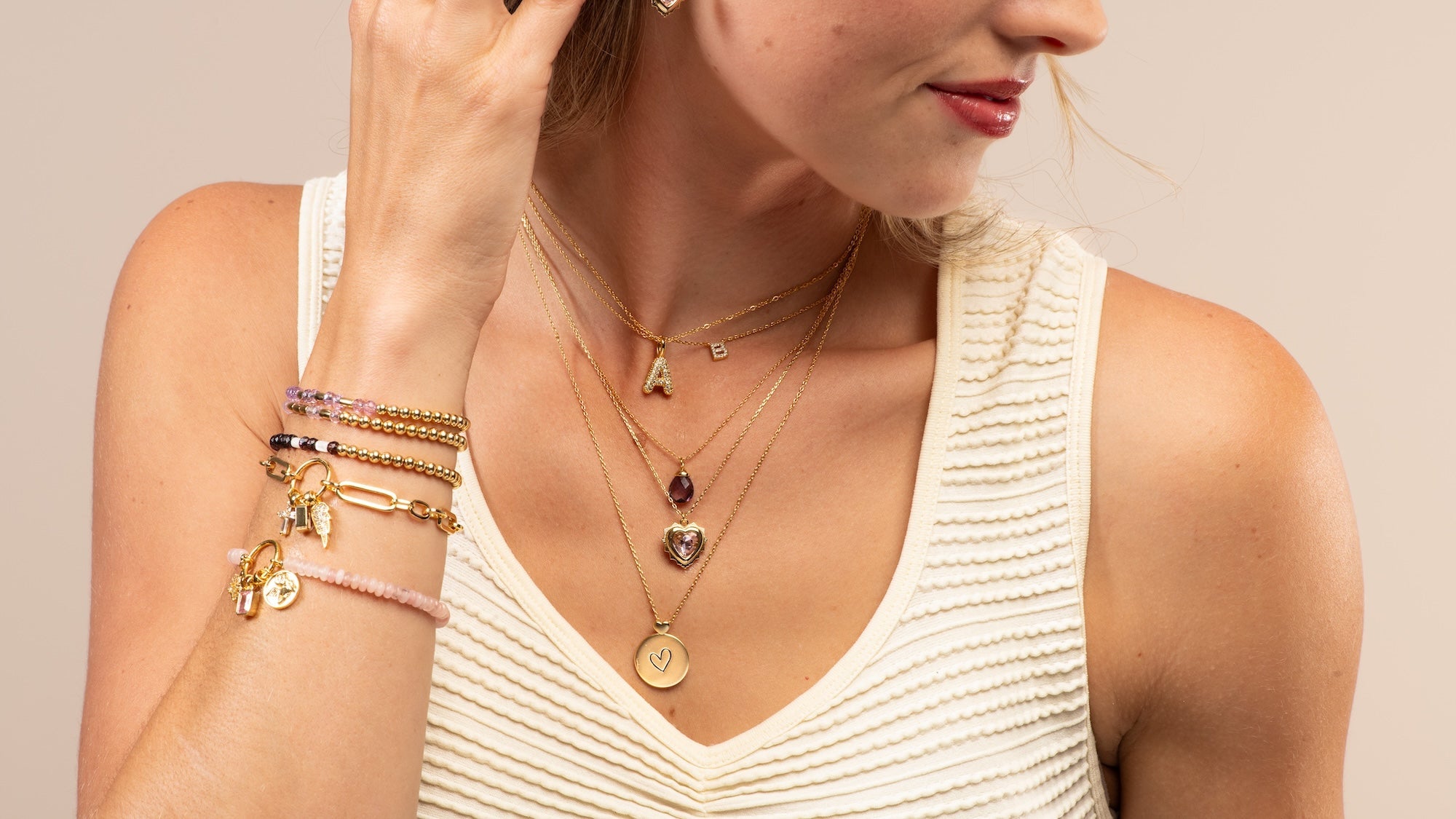 A close-up lifestyle shot of a woman wearing layered gold necklaces and bracelets, including a heart-shaped crystal pendant and initial letters, paired with a cream ribbed tank top against a neutral beige background.