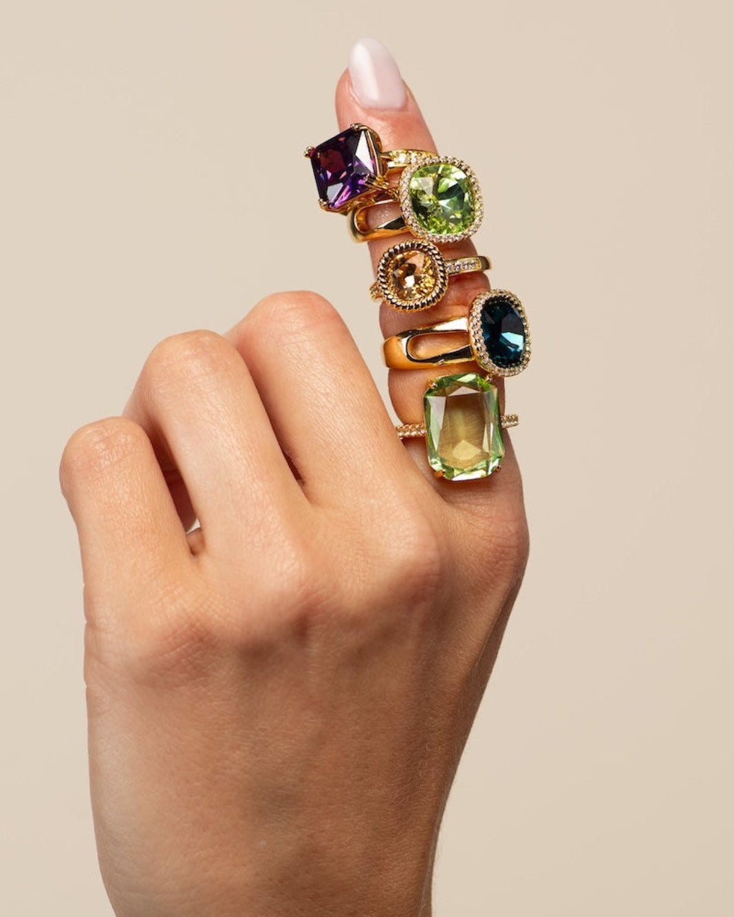 A close-up of a hand wearing a chunky gold ring set with multiple colorful gemstones, including purple, green, and blue stones, against a solid beige background.