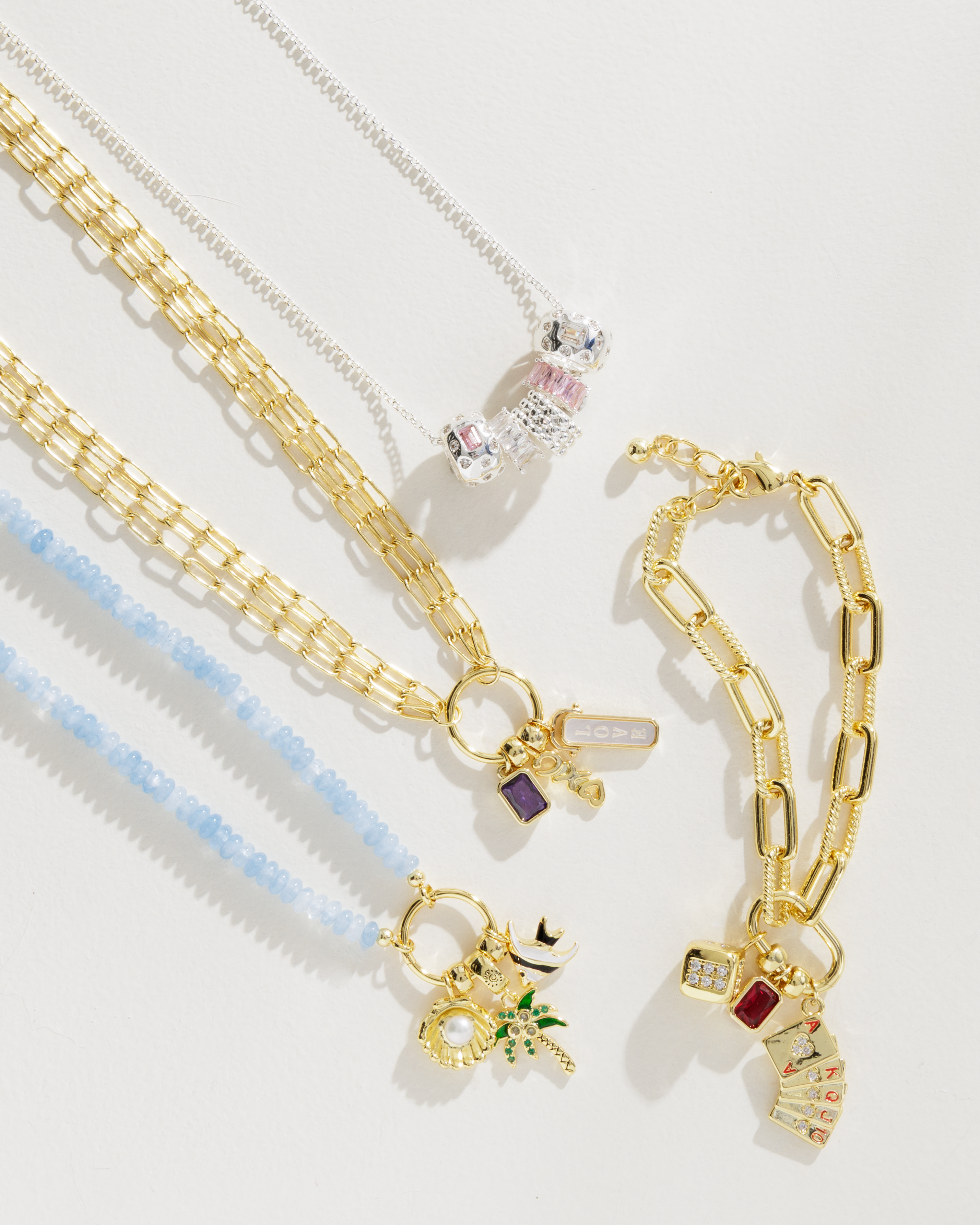 A styled flat-lay of gold-toned jewelry on a white background, featuring a light blue beaded necklace, a gold chain necklace with a small purple and green charm, a silver chain with a pink and white charm, and a gold paperclip bracelet with a small red and white charm.