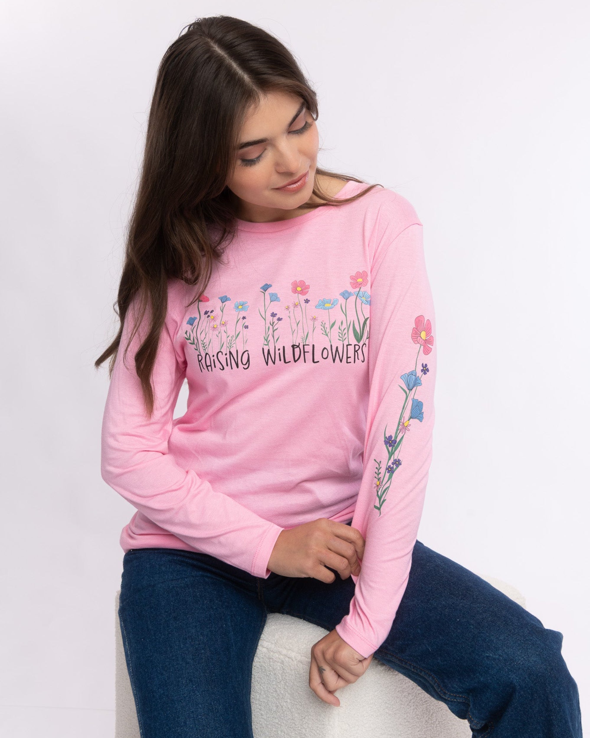 A woman wearing a pink long-sleeve crewneck sweatshirt with 'Blossing Wildflowers' text and floral graphics, paired with dark blue jeans, sitting on a white stool against a white studio background.