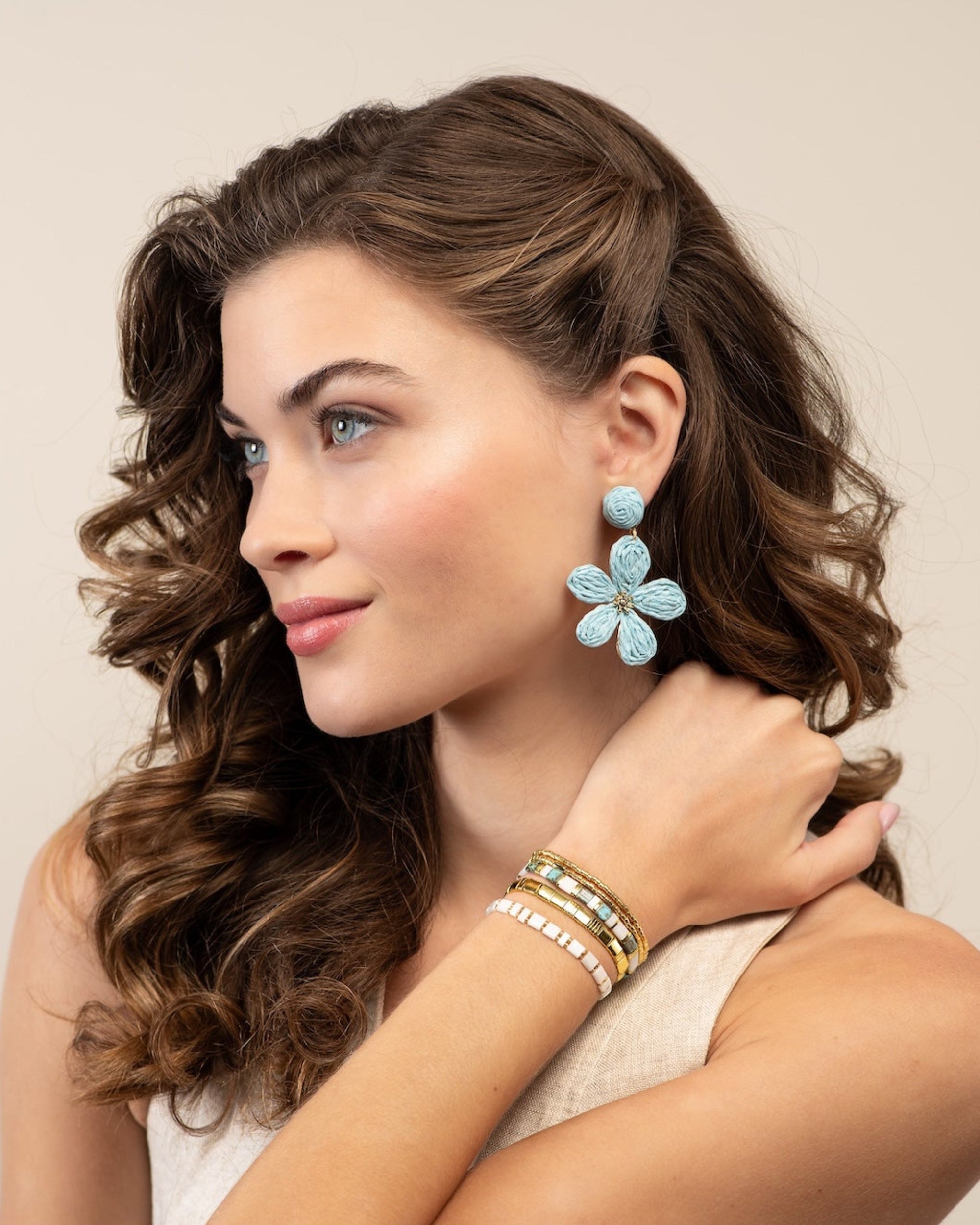 A woman with wavy brown hair is shown in profile wearing light blue flower-shaped drop earrings and stacked gold bracelets against a plain beige studio background.