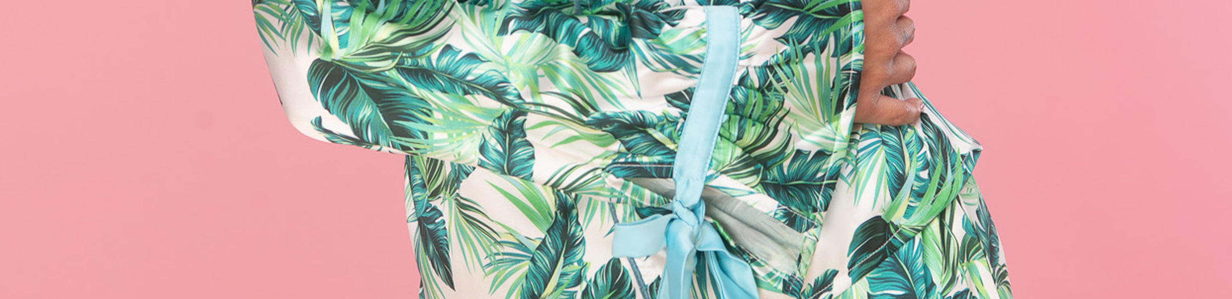 A close-up of a person wearing a tropical leaf print wrap or dress with a light blue ribbon tied at the waist, set against a solid pink background.